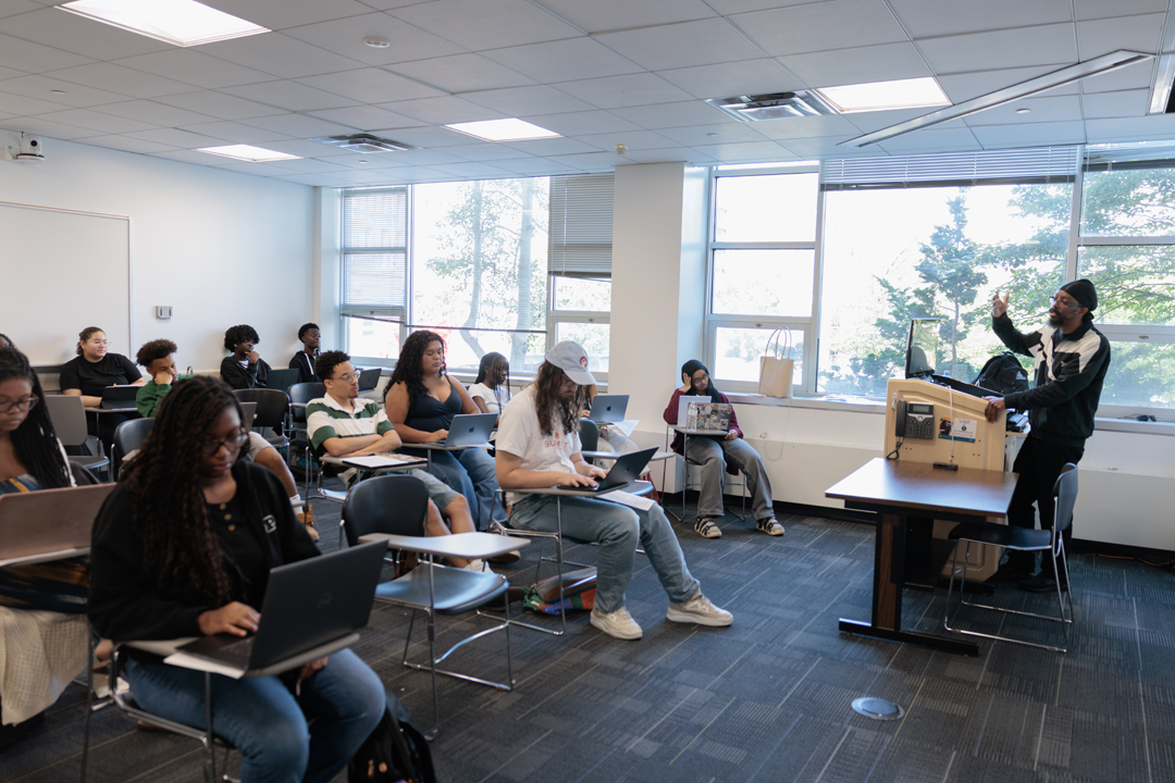 A professor standing and lecturing in a well lit classroom to a full classroom of students seated at desks