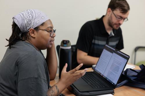 A student talking and gesturing in a small class setting, seated at a table with a professor